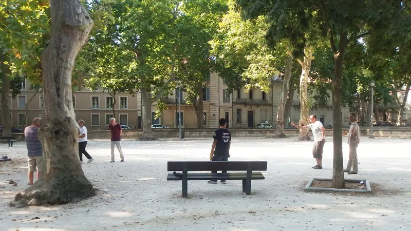Partie de pétanque sur un terrain ombragé par les platanes, avec un banc au premier plan