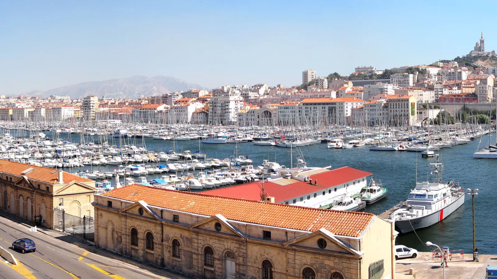Vue panoramique du Vieux-Port de Marseille rempli de voiliers, avec la basilique Notre-Dame de la Garde sur la colline en arrière-plan